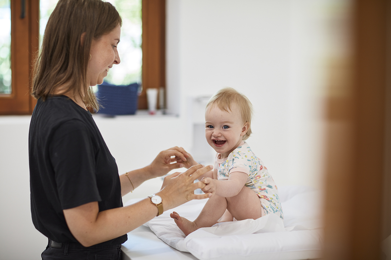 Eine Frau mit langen braunen Haaren lächelt ein glückliches Baby an. Das Baby sitzt auf einer weißen Wickelunterlage und trägt einen bunten Strampler.