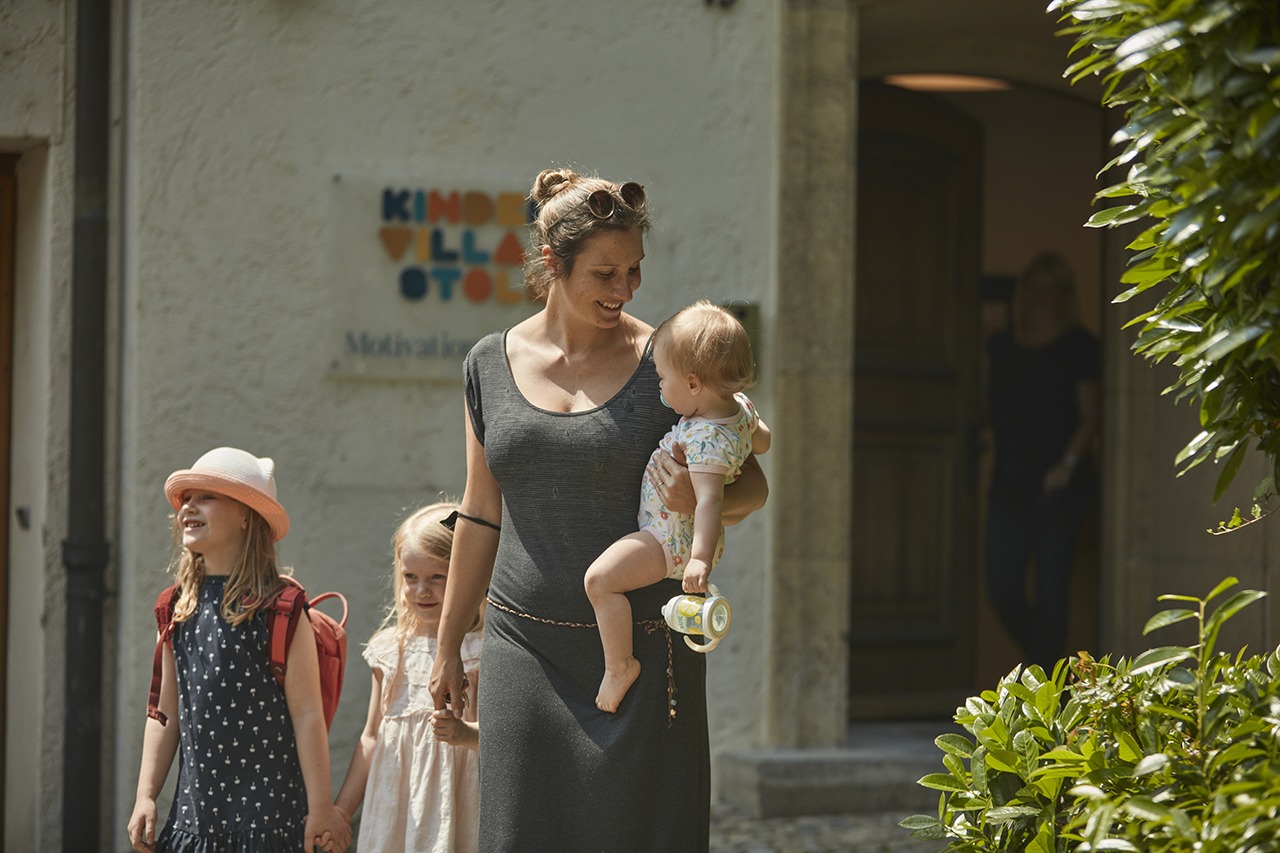 Eine Frau hält ein Kleinkind und geht neben zwei jungen Mädchen in Sommerkleidung und Hüten. Sie sind draußen, in der Nähe von Grünflächen und einem Gebäude mit einem Schild.