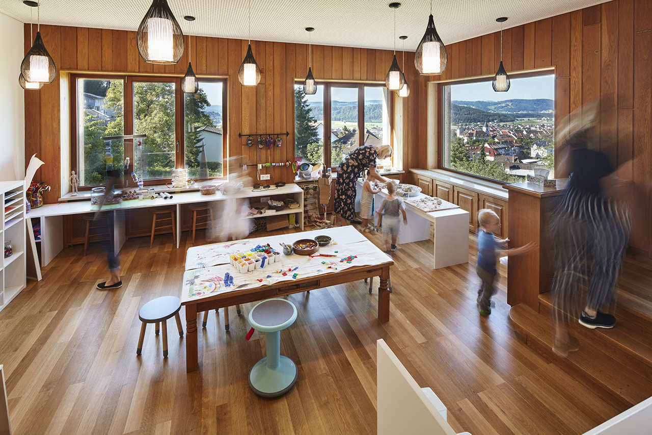 Ein großer, heller Bastelraum mit mehreren Tischen, an denen Kinder und Erwachsene kreativ arbeiten. Der Raum hat große Fenster mit Blick ins Freie. Auf den Tischen liegen Bastelmaterialien, und an den Wänden hängen verschiedene Werkzeuge.