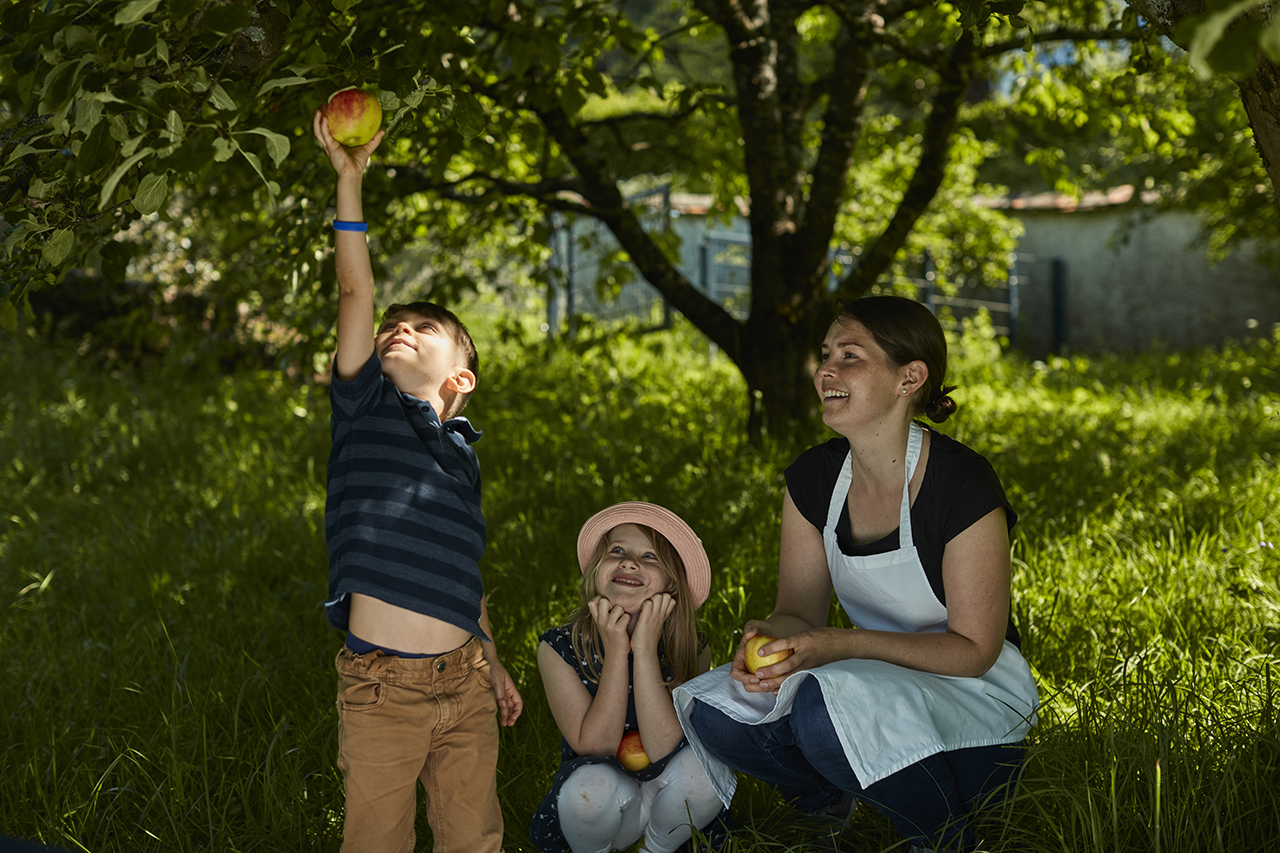 Ein Junge greift nach einem Apfel auf einem niedrigen Ast, während eine Frau und ein Mädchen lächelnd im Gras sitzen und ihn beobachten. Sie befinden sich in einem sonnenbeschienenen Garten mit üppigem grünem Gras und Bäumen.