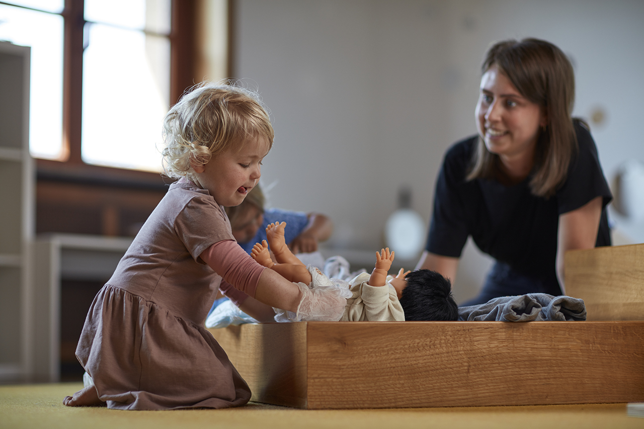 Ein kleines Kind spielt mit Puppen in einer Holzkiste, lächelt und ist fröhlich. Ein Erwachsener, möglicherweise ein Betreuer oder Lehrer, kniet in der Nähe und beobachtet mit warmem Gesichtsausdruck.