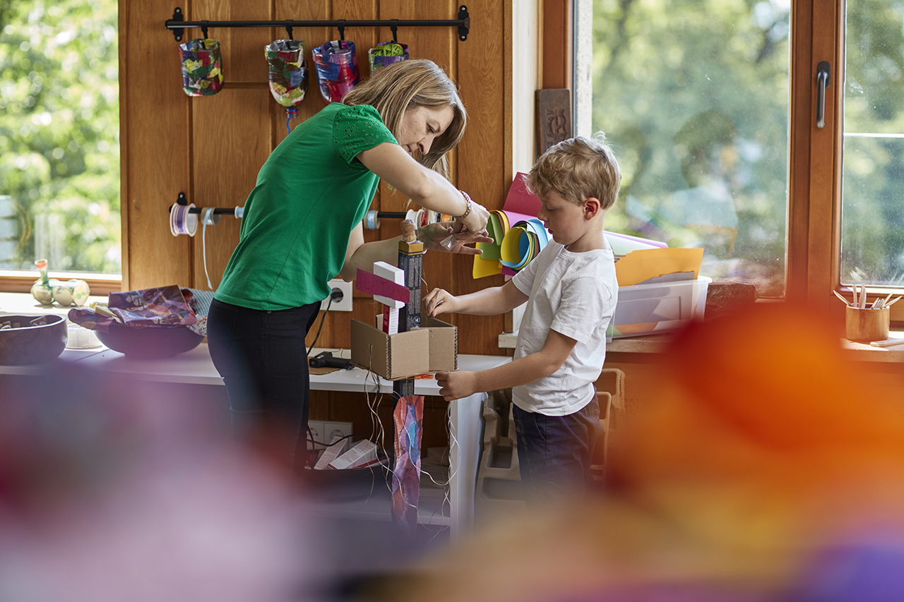 Eine Frau in einem grünen Shirt hilft einem kleinen Jungen beim Basteln. Sie halten gemeinsam eine selbstgebaute Struktur aus Pappe. Im Hintergrund sind bunte Bastelmaterialien und Werkzeuge zu sehen.