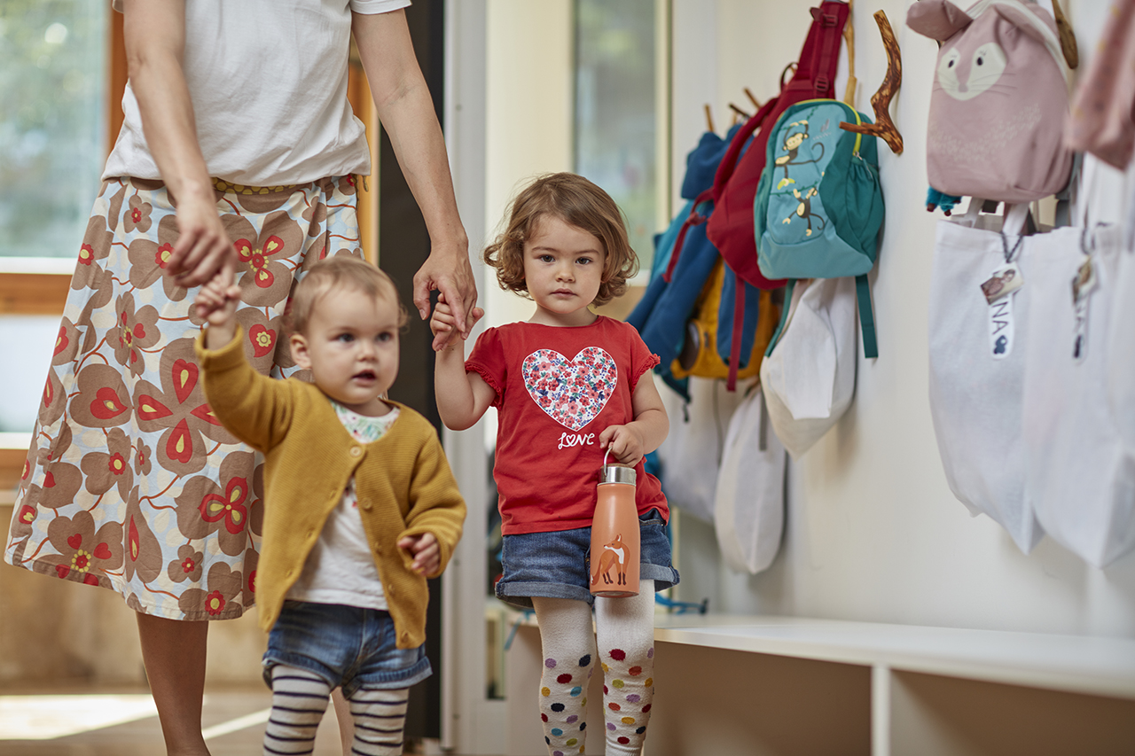 Eine Frau hält die Hände zweier junger Mädchen, die durch einen Flur gehen. Ein Mädchen trägt ein rotes Shirt und gemusterte Leggings und hält eine Wasserflasche. Das andere trägt eine senffarbene Strickjacke und gestreifte Leggings. An der Wand hängen Rucksäcke.