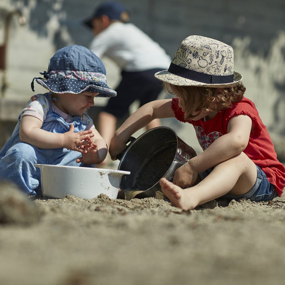 Zwei kleine Kinder spielen an einem Sandstrand. Eines trägt einen blauen Hut und einen Overall, das andere einen Strohhut und ein rotes Hemd. Sie sitzen zusammen, graben und spielen mit Eimern.